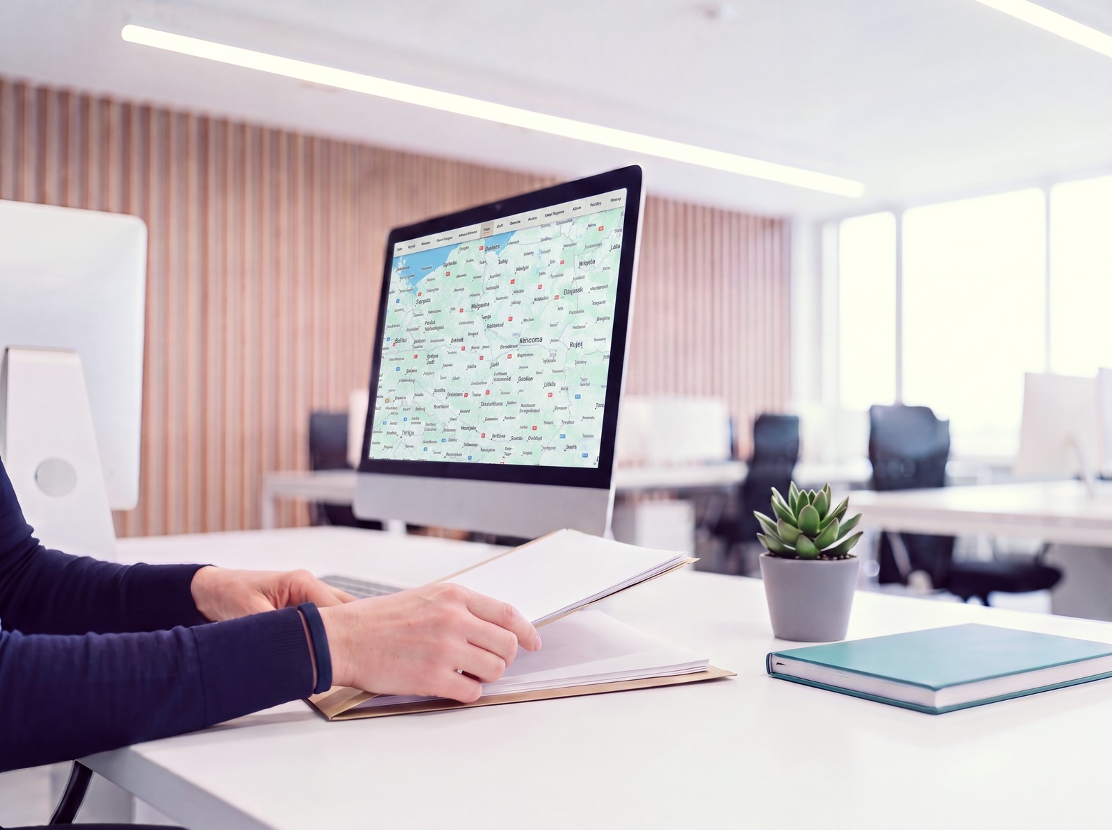 Hands at a folder of blank documents on a white desk, beside a monitor showing a fleet map, the keyboard pushed aside