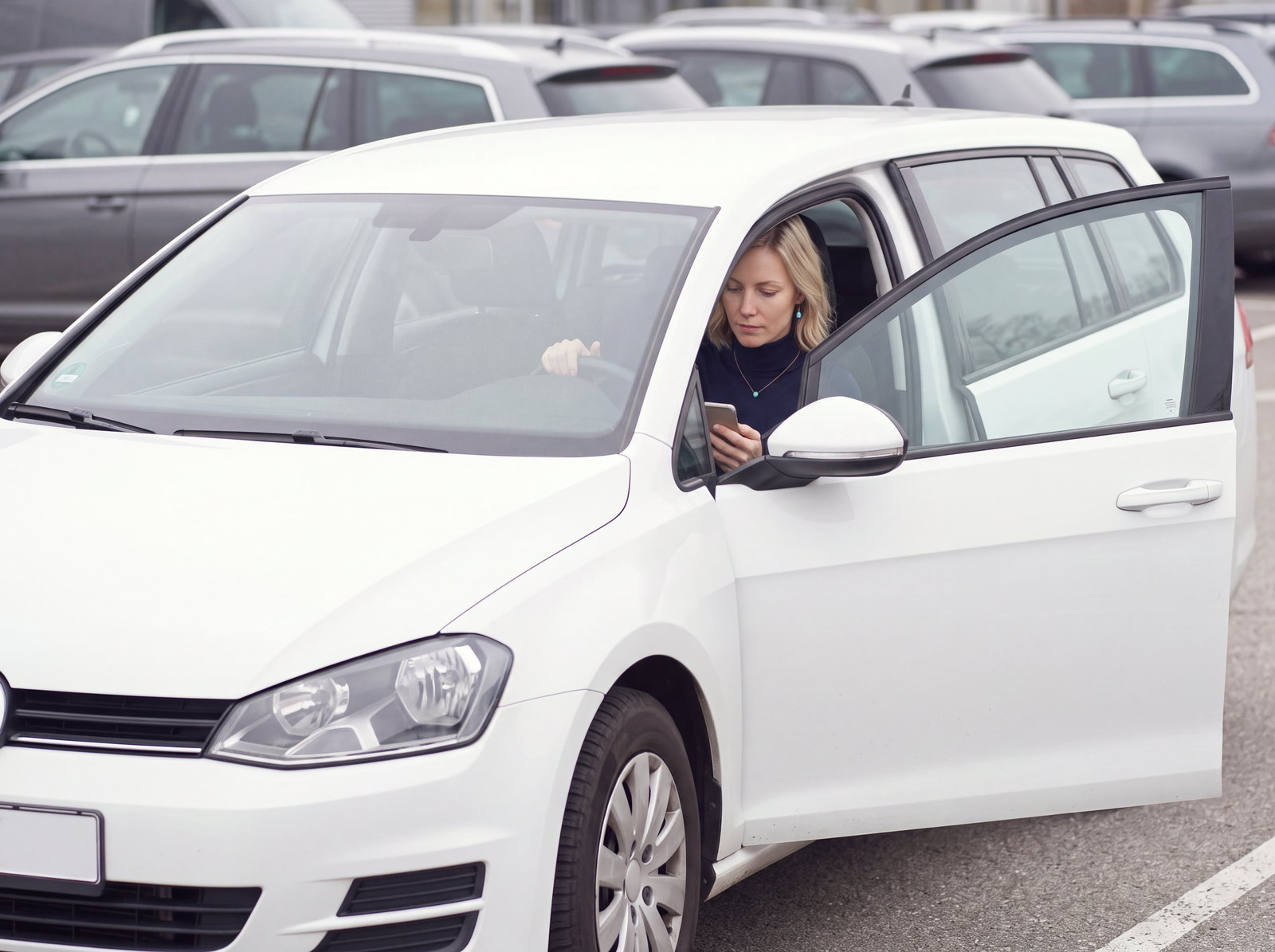 A woman sits behind the wheel of a white estate car with the driver's door open, checking her phone before setting off